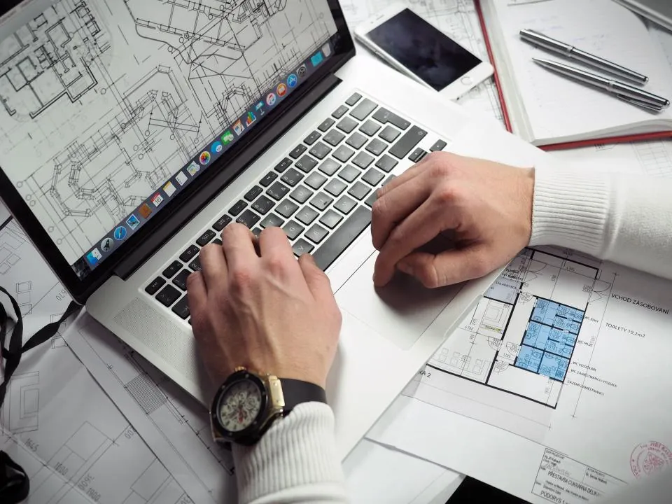 Manos trabajando en un portátil sobre planos y documentos